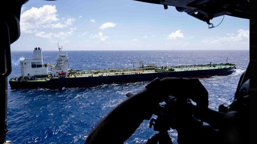 Un gran petrolero oscuro navega en un mar azul, con una estructura blanca visible. El cielo, con nubes blancas, sugiere un día soleado. La imagen fue tomada desde un helicóptero, cuya silueta se aprecia en primer plano.