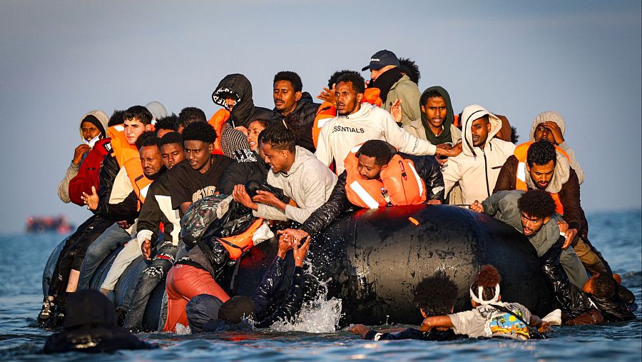 Una embarcación inflable negra, repleta de personas, navega en un mar con olas. Se distinguen individuos con chalecos salvavidas y ropa casual, algunos parecen estar ayudando a otros en el agua.