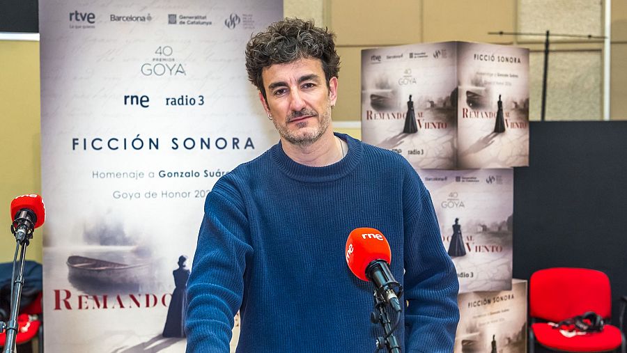 Víctor Clavijo, con barba y jersey azul, se encuentra frente a carteles promocionales que anuncian 