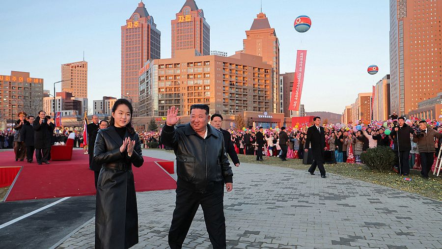 Kim Jong Un y su hija, vestidos con ropa de cuero, saludan a una multitud. Se observa una alfombra roja y globos con los colores de la bandera, con edificios modernos de fondo.