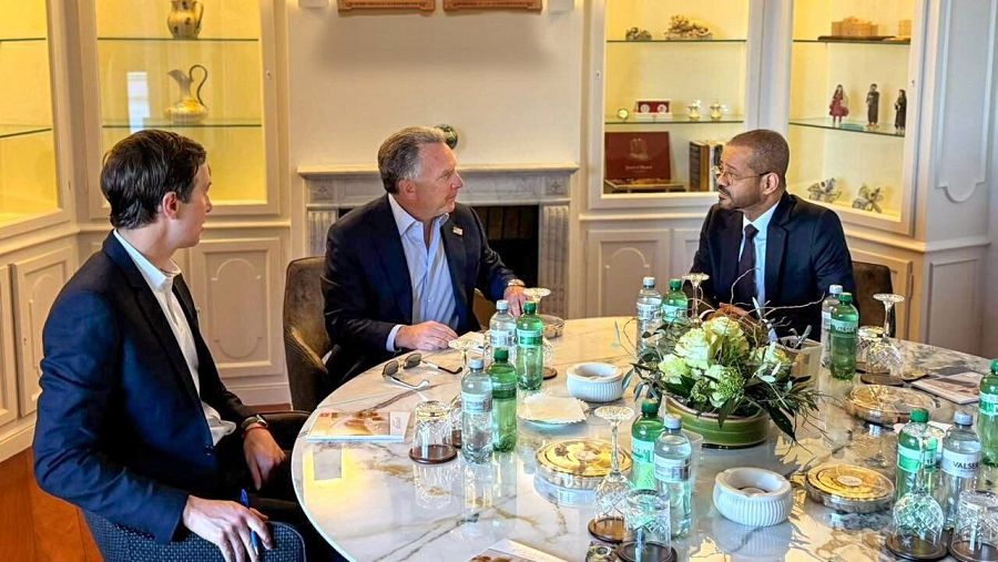 Tres hombres en trajes oscuros, uno con corbata azul, están reunidos en una mesa redonda de mármol con botellas de agua, comida y flores. El ambiente es sofisticado, con armarios y una chimenea decorativa.