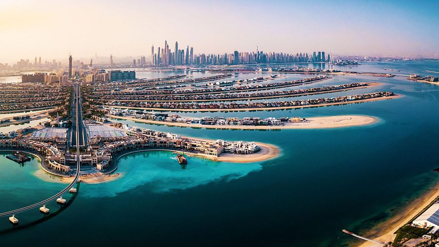 Vista aérea de una ciudad costera con islas artificiales en forma de palmera, densamente edificadas y rodeadas por aguas turquesas. Un horizonte de rascacielos se alza en la distancia, bajo una luz solar intensa.