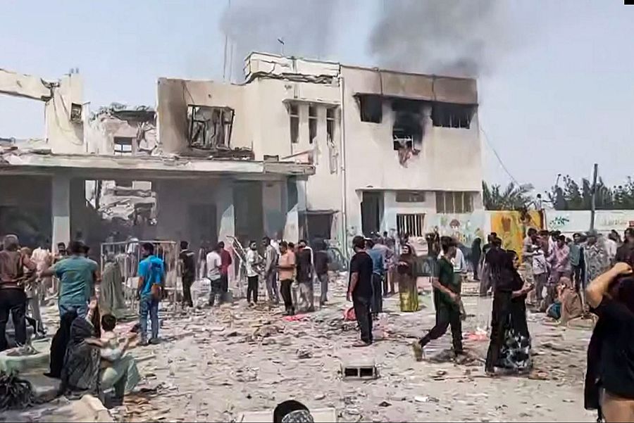 Una escuela en Irán sufre daños significativos tras un ataque. Se observa humo saliendo del edificio con ventanas rotas y escombros en el exterior, donde un grupo de personas se encuentra reunido.