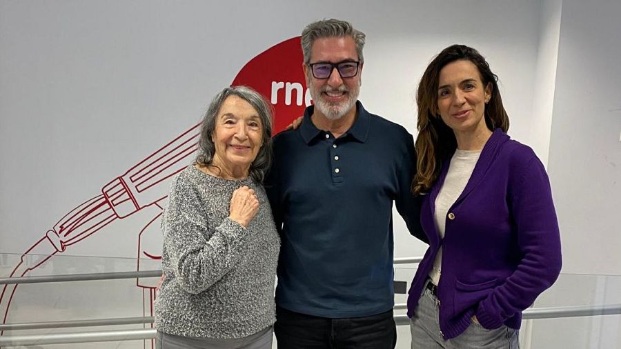 Tres personas posan frente a un fondo blanco con el logo de una emisora de radio. Se distinguen una mujer mayor con suéter gris, un hombre con barba y gafas con un polo azul, y otra mujer con chaqueta morada.