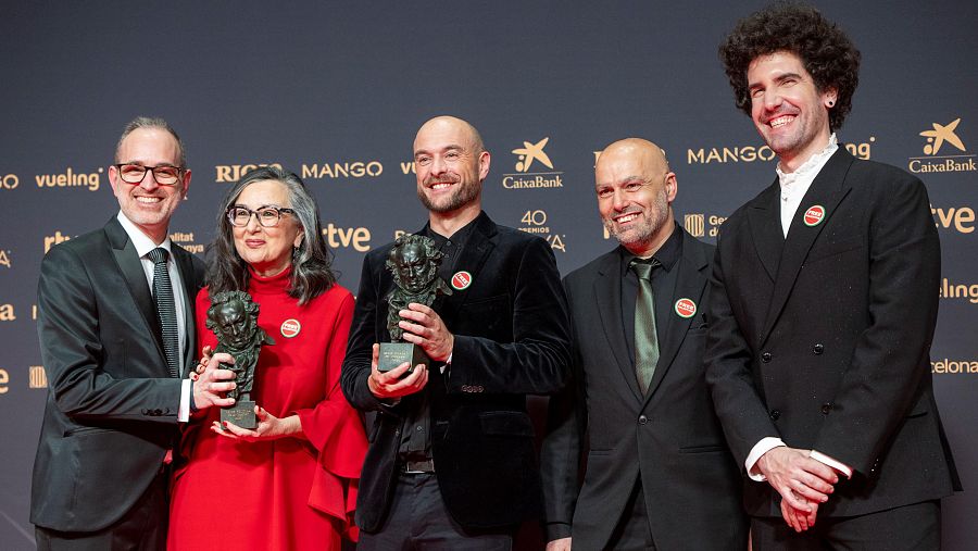Un grupo de personas sonríen y posan con premios, incluyendo a un hombre con gafas y traje negro, una mujer con vestido rojo y un hombre calvo con chaqueta de terciopelo negra.