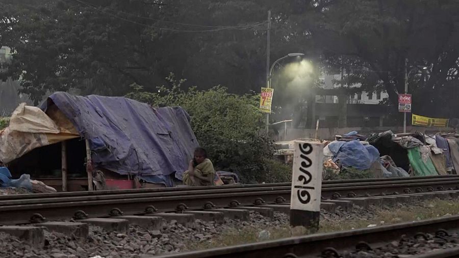 Una vía de tren en Bangladés es el escenario de la imagen, donde se ven refugios improvisados construidos con lonas y otros materiales. Una persona está sentada cerca de las vías, y un poste con texto en bengalí se encuentra en primer plano.