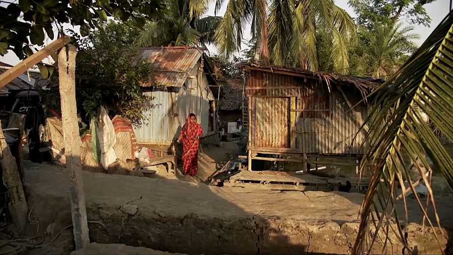 En una aldea, una mujer con un sari rojo y negro camina por un camino de tierra. Se ven casas sencillas y vegetación tropical, sugiriendo un clima cálido y un terreno seco.