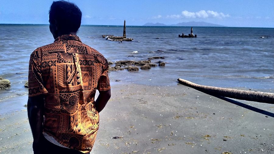 Un hombre con una camisa estampada observa el mar desde una playa de arena gris, donde se aprecian estructuras que emergen del agua, posiblemente de un cementerio. Al fondo, se distinguen montañas bajo un cielo azul.