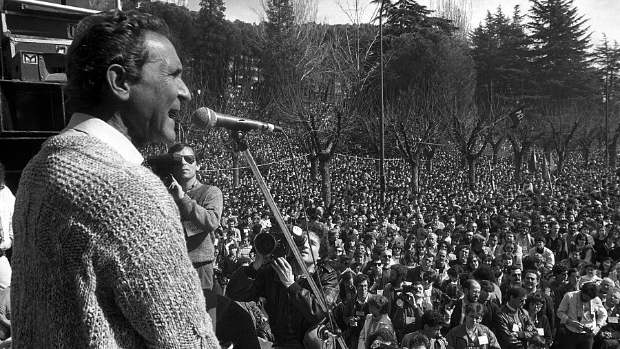 Antonio Gala, de perfil y con un micrófono, habla ante una multitud en un evento al aire libre, posiblemente en 1986. Viste un jersey de punto grueso y está rodeado de personas y árboles.
