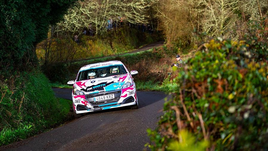 En la imagen puede observarse un coche de color blanco de rally que está tomando una curva en la carretera. En su interior hay dos mujeres.