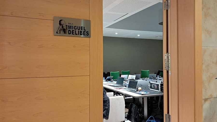 Una puerta entreabierta muestra una sala de reuniones con una mesa grande y sillas. Sobre la mesa, se observan ordenadores portátiles y equipos electrónicos. La puerta tiene una placa con un nombre y una imagen.