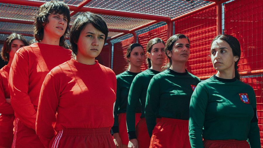 Un grupo de mujeres jóvenes, vestidas con camisetas y faldas, posan frente a una estructura metálica roja. Algunas llevan camisetas rojas y otras verdes, todas mirando hacia el frente con seriedad.