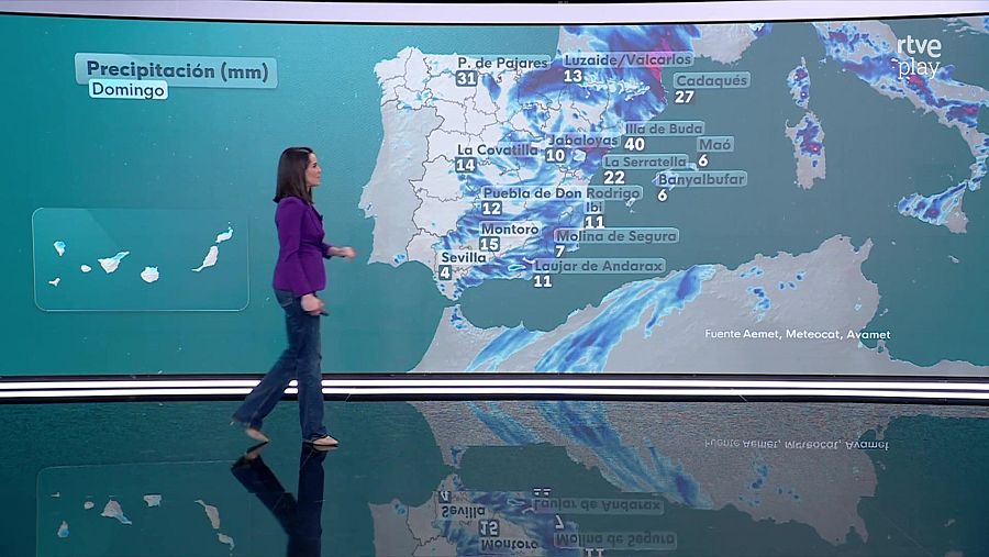 Un mapa de España y las Islas Canarias muestra las precipitaciones del domingo en milímetros. Una mujer camina frente al mapa, vestida con chaqueta morada, vaqueros y zapatos marrones. Se indican cantidades de precipitación en varias localidades.