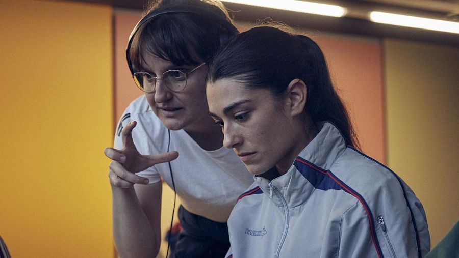Dos mujeres, una con gafas y auriculares y otra con coleta, interactúan frente a una pared con paneles de colores, posiblemente concentradas en algo fuera de la imagen.