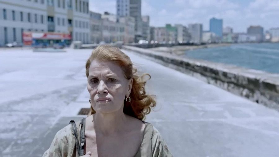 Una mujer de mediana edad, con cabello rojizo y piel clara, posa con pendientes dorados y una blusa clara, llevando un bolso. El fondo muestra edificios antiguos y el mar, posiblemente en una ciudad costera.