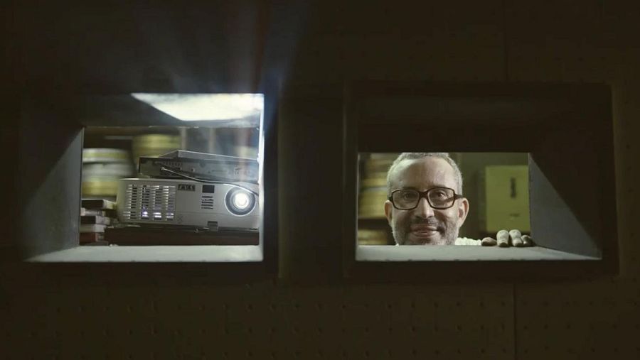 Un fotograma de 'Neurótica anónima' muestra a un hombre con gafas sonriendo a la cámara a través de una abertura en la pared, con un proyector y latas de película visibles en otra abertura.