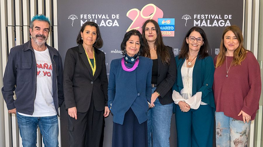 Un grupo de seis personas posa para una foto. Entre ellos, se distinguen individuos con vestimentas variadas, desde trajes formales hasta ropa casual, frente a un fondo con logotipos de un festival.
