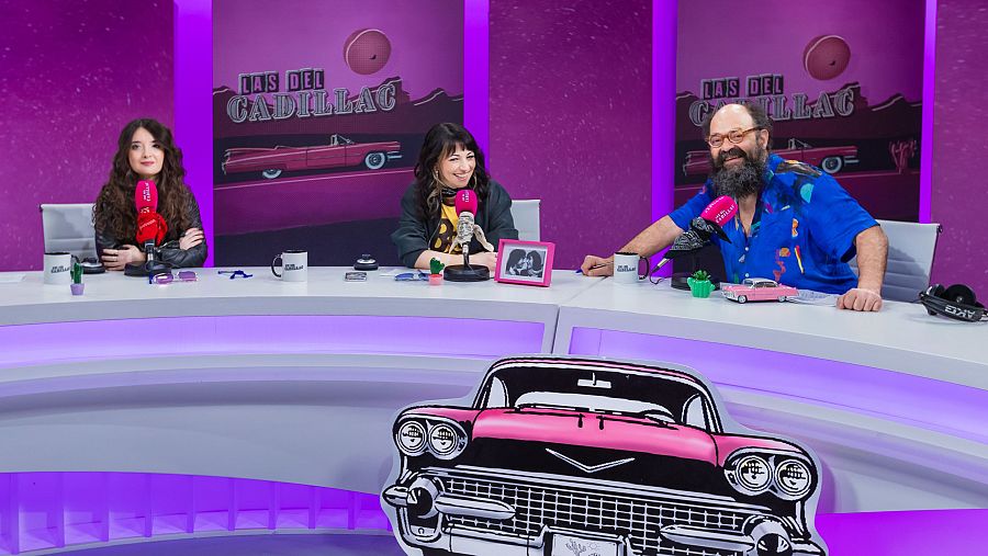 Tres personas, María Canet, Raquel Elices e Ignatius Farray, están sentadas en una mesa frente a micrófonos rojos, con carteles que anuncian 