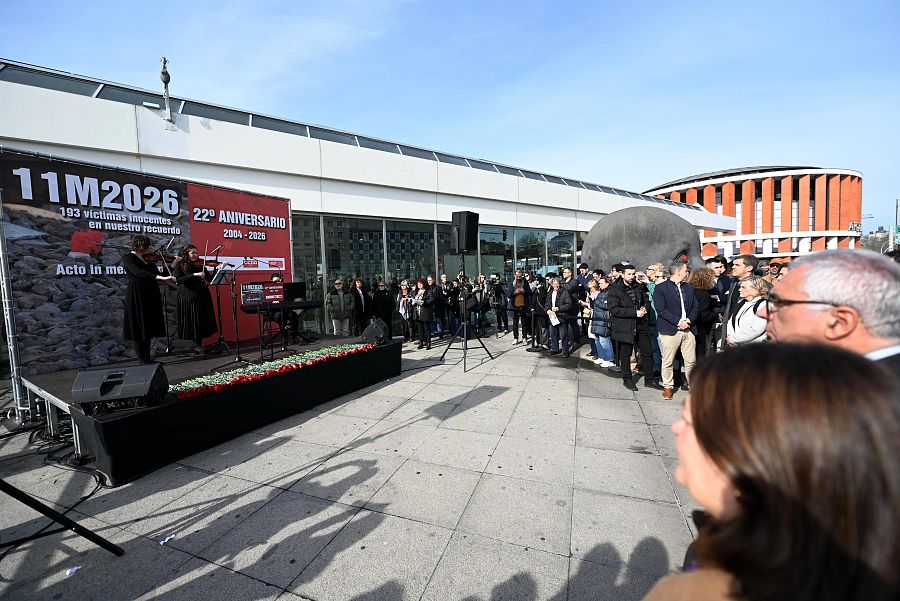 Un acto solemne conmemora el 22º aniversario de los atentados del 11M en la estación de Atocha. Músicos tocan en un escenario frente a una audiencia, con flores y una pancarta en memoria de las víctimas.