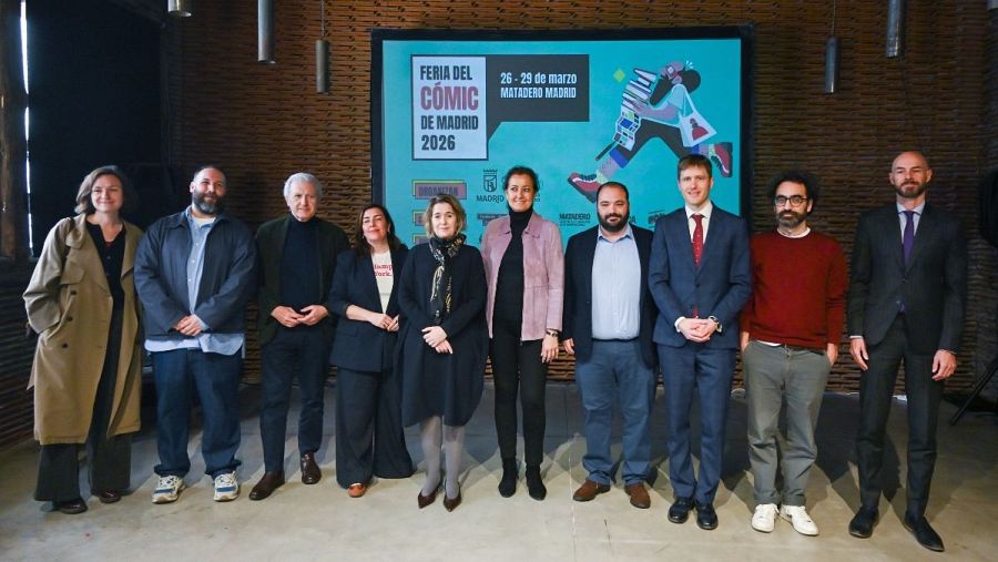 Un grupo de personas posa frente a un cartel promocional. El cartel anuncia un evento con una ilustración de una figura corriendo con libros. Los individuos visten ropa variada, desde casual hasta formal.