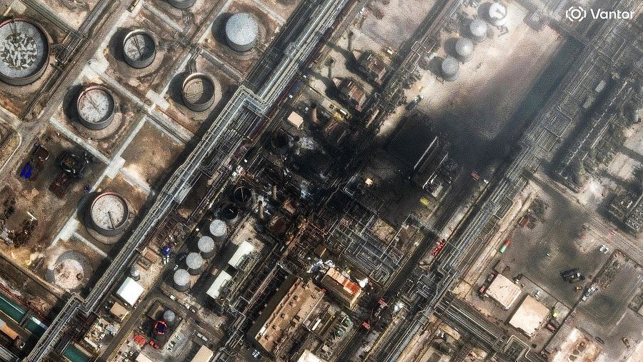 Vista aérea de una refinería de petróleo con daños visibles, incluyendo tanques de almacenamiento y una red de tuberías. Se aprecia hollín y evidencia de fuego en una parte considerable de la instalación, indicando un incidente de gran magnitud.