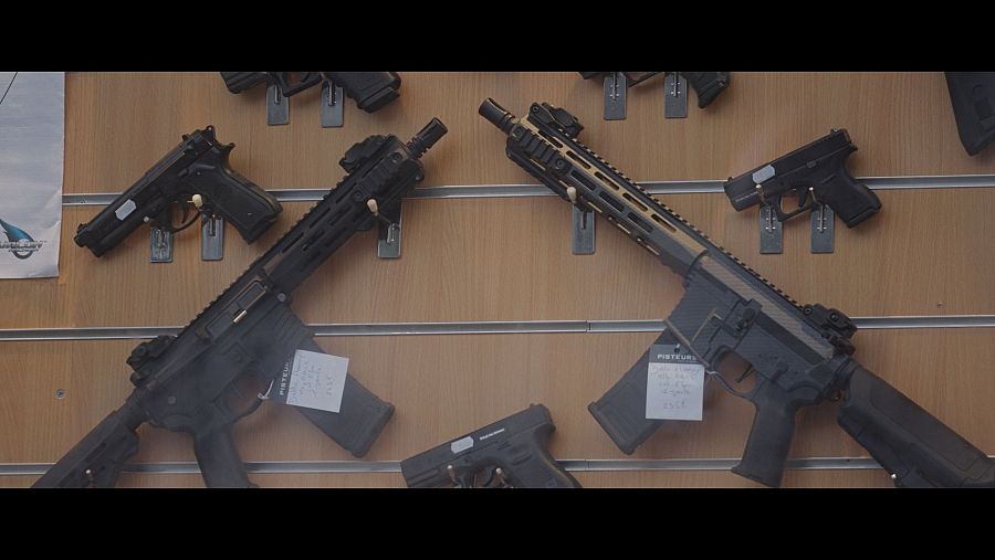 En una pared de madera se exhiben diversas armas de fuego, como rifles de asalto y pistolas, sujetas con soportes metálicos. Se distinguen armas de color negro y otras con detalles en arena, cada una con una etiqueta.