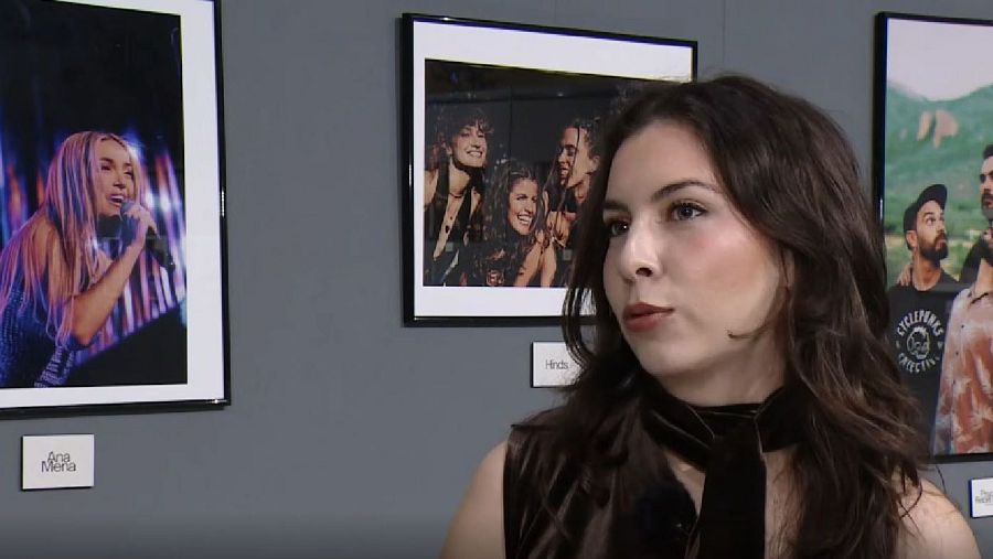 Una mujer con cabello castaño y un vestido oscuro posa frente a una pared gris donde se exhiben tres fotografías enmarcadas, mostrando a diferentes personas.