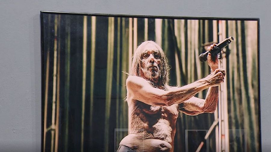 En la imagen, Iggy Pop, con el torso desnudo y cabello canoso, canta en un escenario. Sostiene un micrófono y se encuentra frente a cortinas verticales oscuras y doradas.
