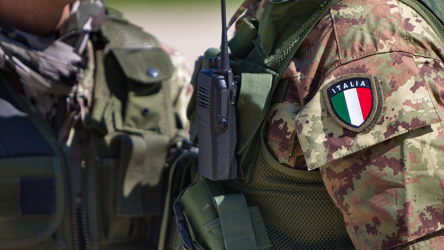 Dos militares italianos, vestidos con uniformes de camuflaje y chalecos tácticos, posan para la foto. Uno de ellos porta una radio y un parche con la bandera italiana.