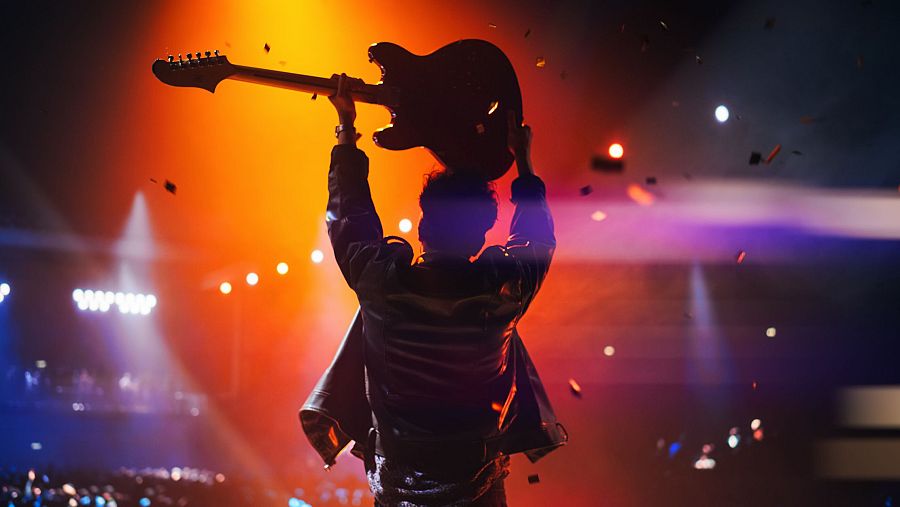 En un escenario iluminado con luces de colores, un artista con una chaqueta brillante levanta una guitarra sobre su cabeza, mientras el público observa el espectáculo y cae confeti.