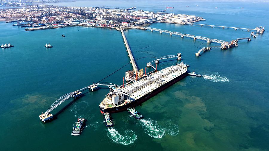 Un petrolero es guiado hacia un muelle en una terminal petrolera, con varios barcos pequeños asistiendo en la maniobra. Se aprecia una ciudad al fondo y diversas estructuras portuarias.