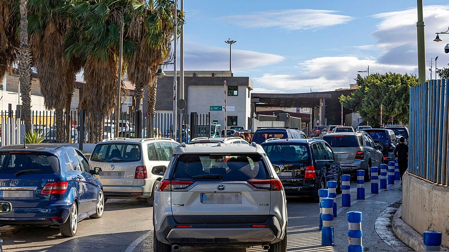 Una larga hilera de coches de distintos colores avanza lentamente hacia el control fronterizo, con edificios y vegetación a los lados de la carretera.