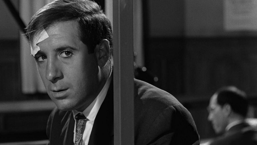 Alfredo Landa, vestido con traje y corbata, muestra una expresión de sorpresa con una tirita en la frente. Parte de su cuerpo se oculta tras una columna, mientras que al fondo se vislumbra otra figura.