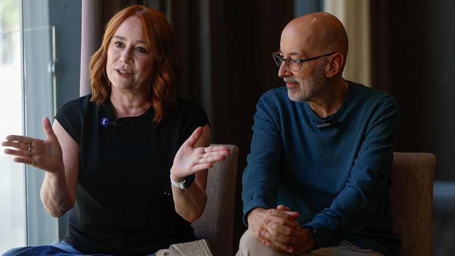 Gracia Querejeta y Miguel Olid, ambos con micrófonos de solapa, aparecen sentados. Ella, con camiseta negra y reloj, gesticula; él, con jersey azul y gafas, escucha con las manos entrelazadas.