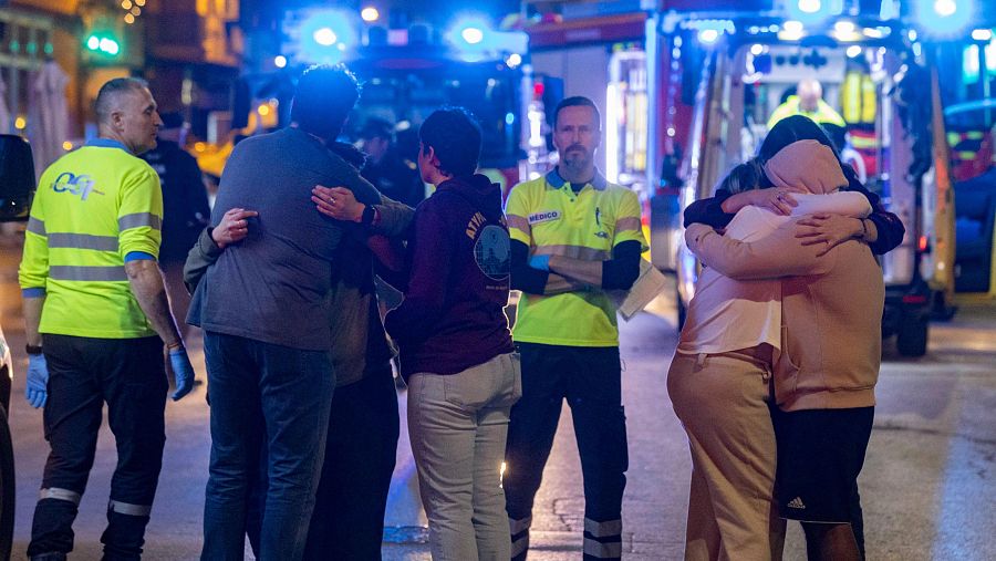 Un grupo de personas se abrazan consolándose mientras miembros del equipo de emergencias continúan sus trabajos