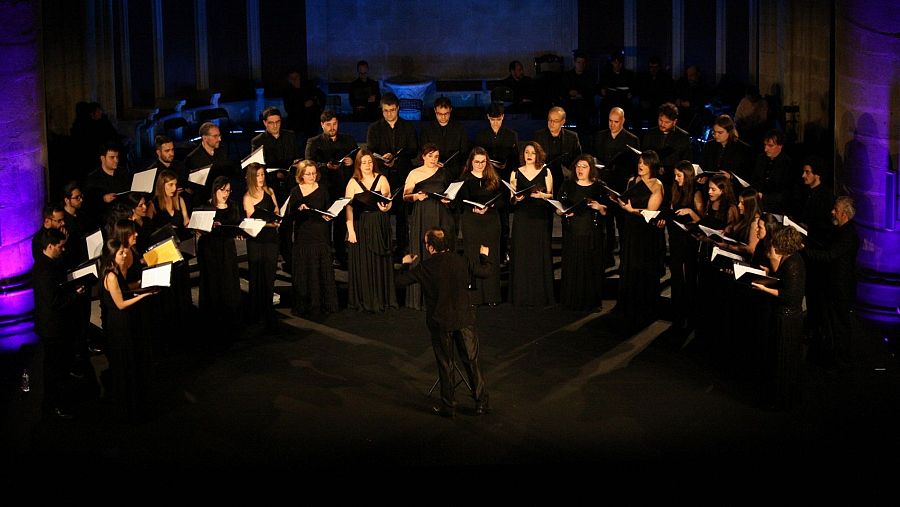 Un numeroso coro, vestido de negro, canta bajo la dirección de un director de orquesta. Los cantantes sostienen partituras en un espacio con arquitectura clásica, iluminado tenuemente.