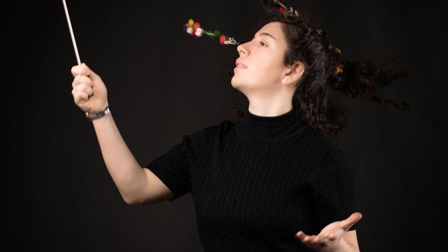 Una mujer joven, con cabello oscuro y rizado decorado con flores, dirige una orquesta. Viste un suéter negro y sostiene una batuta blanca, con el fondo oscuro.