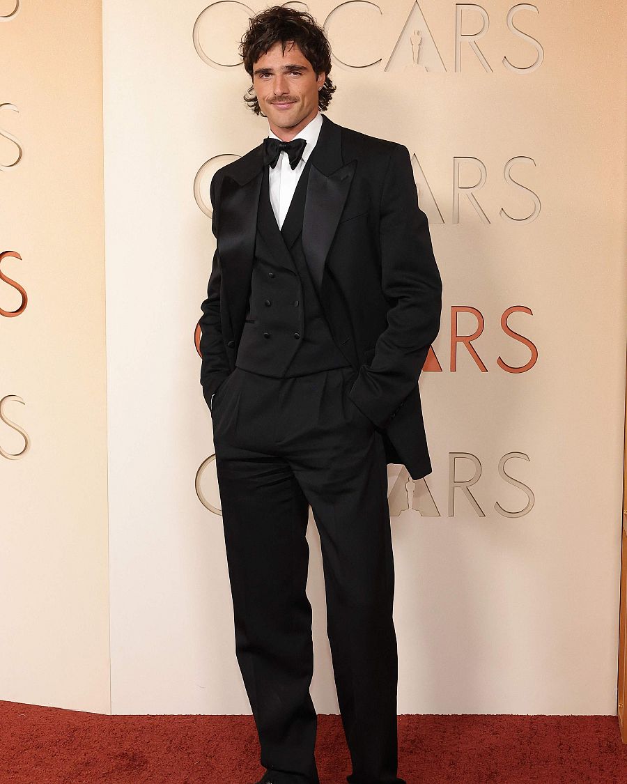 En la alfombra roja, un hombre con traje negro y bigote posa frente a una pared con la palabra 