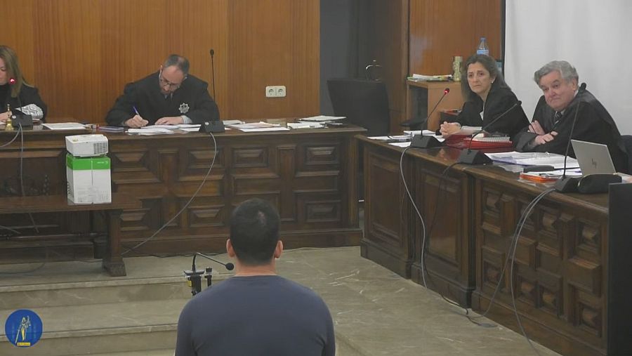 En una sala de juicios, se observa a una persona de espaldas con una camiseta oscura. Detrás de un escritorio, cuatro individuos con togas, posiblemente jueces, están presentes, con micrófonos, una computadora y papeles.