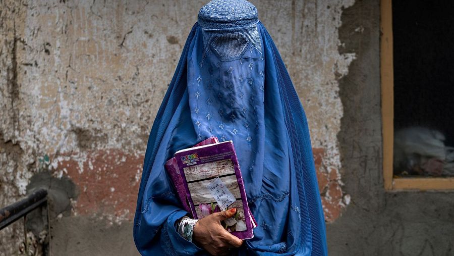 Una mujer con burka azul, con detalles bordados, sostiene dos cuadernos morados. En uno de ellos se lee una nota. Se aprecia una pulsera plateada en su muñeca, y el fondo es una pared con una ventana.