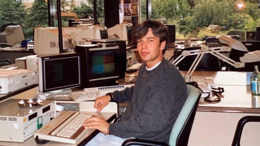 Óscar Gutiérrez, ex redactor de teletexto, está sentado frente a un teclado en una oficina con múltiples ordenadores y teclados, posiblemente trabajando. Viste un jersey gris y una camisa blanca.