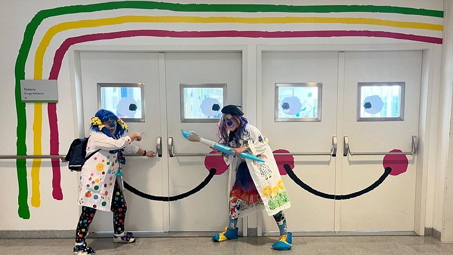 Dos payasas, la Doctora Lunarete y la Doctora Pintorina, posan frente a unas puertas decoradas con dibujos. Una de ellas señala hacia la puerta, mientras que la otra sostiene un pincel inflable. El entorno incluye un arcoíris pintado y un cartel.