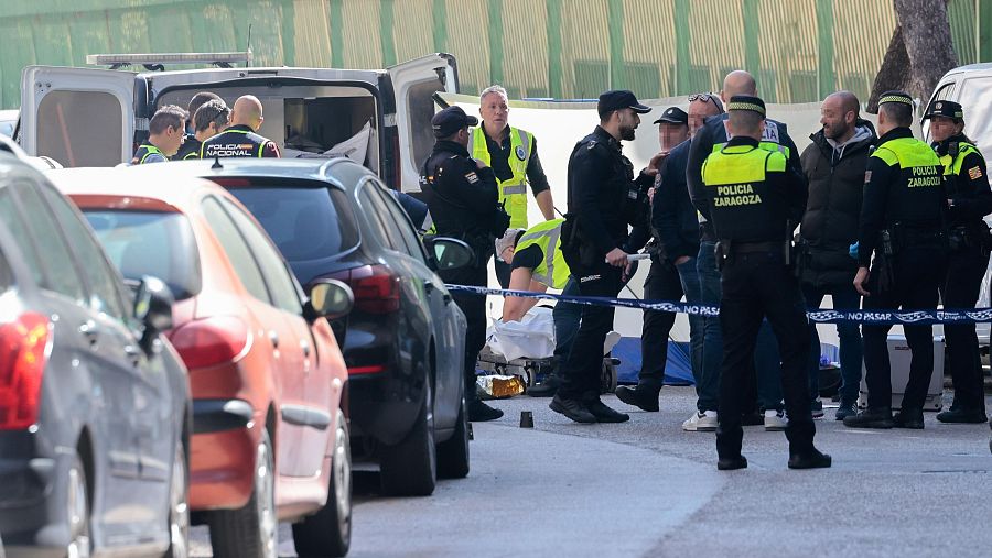 Una escena de investigación policial en la vía pública muestra coches estacionados, agentes de policía y personal sanitario atendiendo a una persona. Una cinta policial restringe el acceso a la zona.