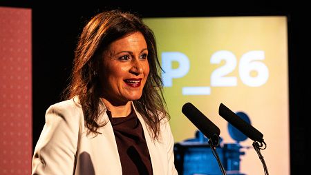Sara Martín Olmo, con cabello castaño oscuro y labios rojos, sonríe frente a dos micrófonos. Viste una blusa oscura y una chaqueta blanca, con un cartel amarillo y texto blanco al fondo.