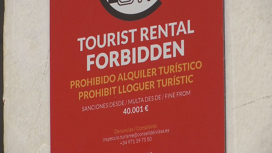 Un cartel rojo advierte sobre la prohibición del alquiler turístico, con textos en inglés, español y catalán, y detalles sobre las sanciones económicas, además de información de contacto para denuncias.