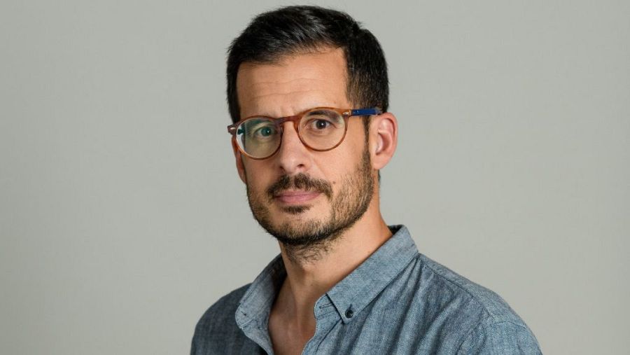 José Luis Sastre, con gafas de montura redonda y camisa azul claro, posa frente a un fondo gris claro.
