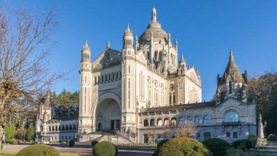 Una fotografía de la Basílica de Santa Teresa de Lisieux, con su imponente estructura de piedra clara y cúpula central. Se observan detalles arquitectónicos como arcos y ventanas, junto a zonas verdes en primer plano.