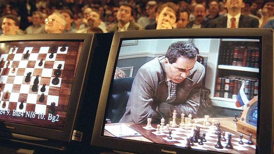 Dos monitores en primer plano: uno muestra un tablero de ajedrez y otro a Garry Kasparov apoyado sobre la mesa; al fondo, el público observa siguiendo la partida en una pantalla grande.