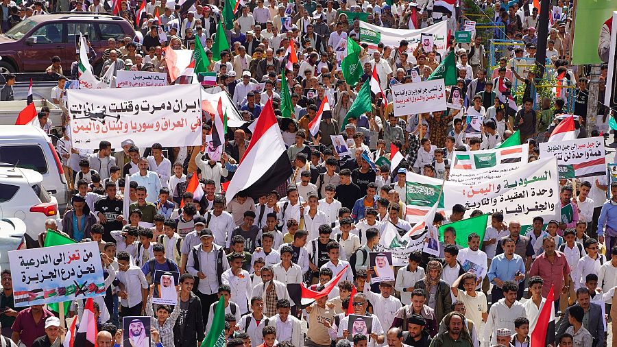 Una manifestación en una calle muestra a una multitud con pancartas y banderas, incluyendo las de Yemen, Arabia Saudita e Irán. Los carteles en árabe expresan mensajes políticos y algunas personas exhiben fotos del príncipe heredero saudí.