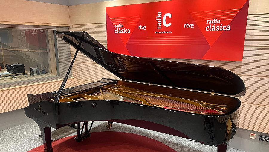 En un estudio de radio, un piano de cola negro Steinway & Sons se encuentra frente al logotipo de Radio Clásica, con su diseño distintivo. El entorno se completa con paredes claras, una alfombra roja y una ventana.